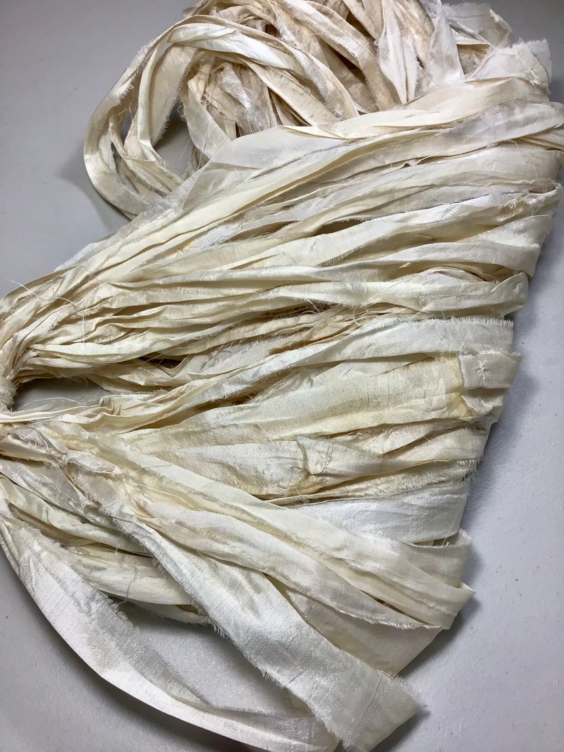 May include: Cream-colored silk ribbon in a tangled bundle. The ribbon has a slightly textured appearance with frayed edges, suggesting a handmade or vintage quality. The color is a soft, off-white, with subtle variations in tone.