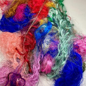 May include: A pile of colorful, textured fiber in shades of red, blue, pink, green, and gold. The fibers appear to be loosely spun and have a slightly shiny appearance. The image is a close-up, highlighting the variety of colors and textures.