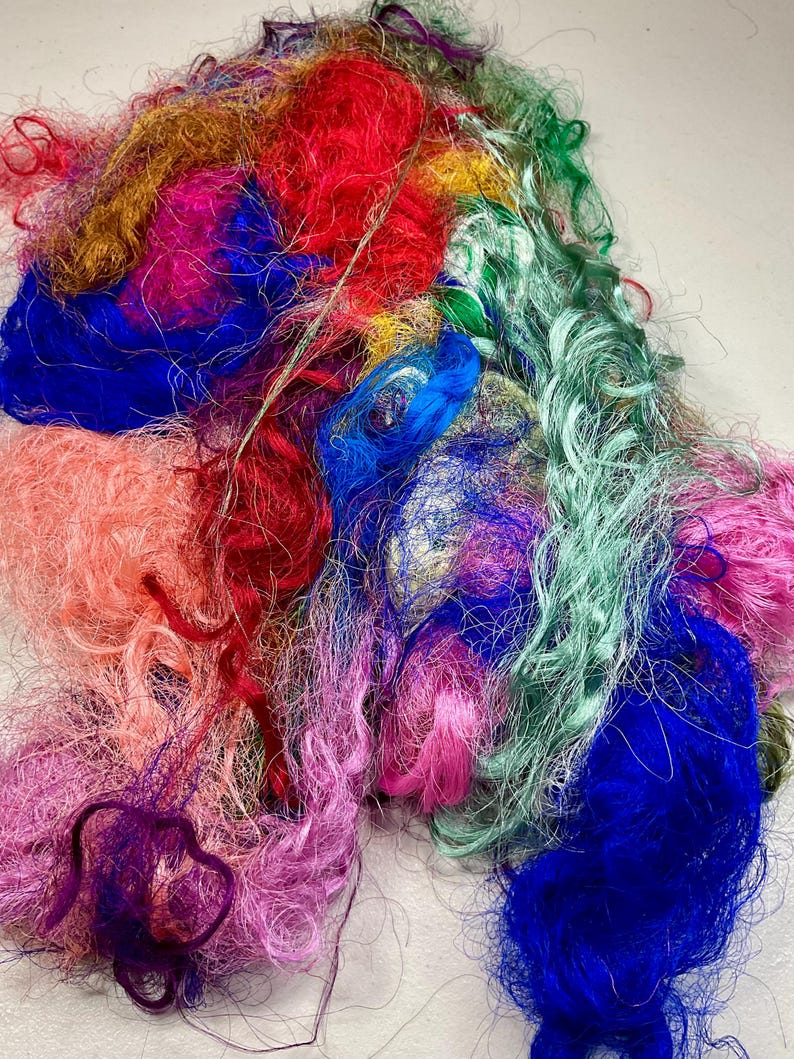 May include: A pile of colorful, textured fiber in shades of red, blue, pink, green, and gold. The fibers appear to be loosely spun and have a slightly shiny appearance. The image is a close-up, highlighting the variety of colors and textures.