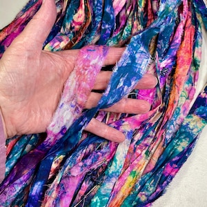 May include: A bundle of colorful, hand-dyed fabric strips in shades of pink, blue, and teal. The fabric has a textured, frayed edge and a tie-dye appearance. The strips are held in a hand, showcasing the vibrant colors and unique patterns.
