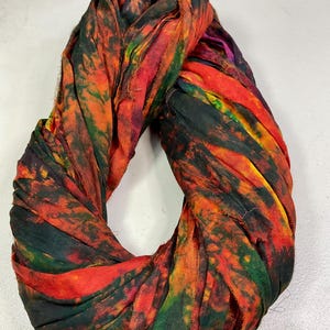 May include: A colorful, hand-dyed scarf in a circular shape. The scarf features a vibrant mix of colors, including red, orange, green, and black. The fabric appears to be soft and flowing, with a textured, tie-dye pattern. The scarf is a fashion accessory.