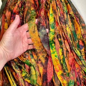 May include: Close-up of colorful fabric strips in shades of orange, yellow, green, and brown. The strips have a textured, hand-dyed appearance, with visible variations in color and pattern. The fabric is held by a hand.