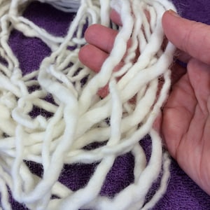 May include: A pile of thick, off-white yarn, with a soft, textured appearance. The yarn is held in a hand, showing the individual strands. The background is a solid purple color.
