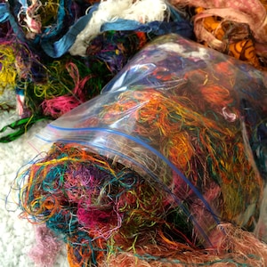 May include: A close-up shot of a collection of colorful fabric scraps. The scraps are a mix of textures and colors, including blue, orange, pink, and yellow. Some scraps are in a clear plastic bag, while others are loose. The image is well-lit and the colors are vibrant.