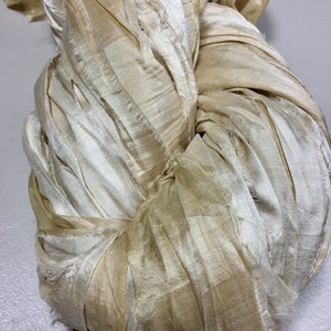 May include: A close-up of a bundle of ivory-colored silk ribbon. The ribbon is loosely twisted and knotted, with a slightly frayed texture. The color varies from a pale cream to a light tan.