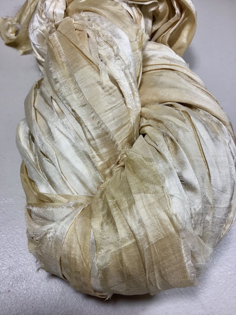 May include: A close-up of a bundle of ivory-colored silk ribbon. The ribbon is loosely twisted and knotted, with a slightly frayed texture. The color varies from a pale cream to a light tan.