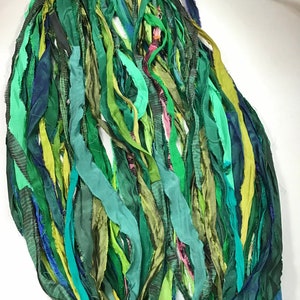 May include: An abstract arrangement of long, flowing fabric strips in various shades of green, teal, and blue. The strips are layered and textured, creating a sense of depth and movement. Some strips have subtle patterns.