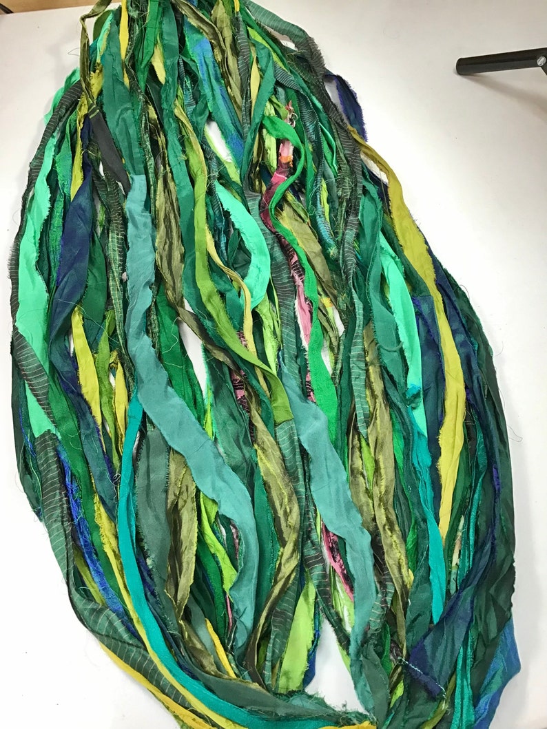 May include: An abstract arrangement of long, flowing fabric strips in various shades of green, teal, and blue. The strips are layered and textured, creating a sense of depth and movement. Some strips have subtle patterns.