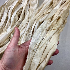 May include: Cream-colored silk ribbon, with a slightly frayed edge, is held in a hand. The ribbon has a soft, textured appearance and is arranged in loose folds. The color is a warm, off-white, suggesting a natural dye. This ribbon is suitable for crafts and embellishments.