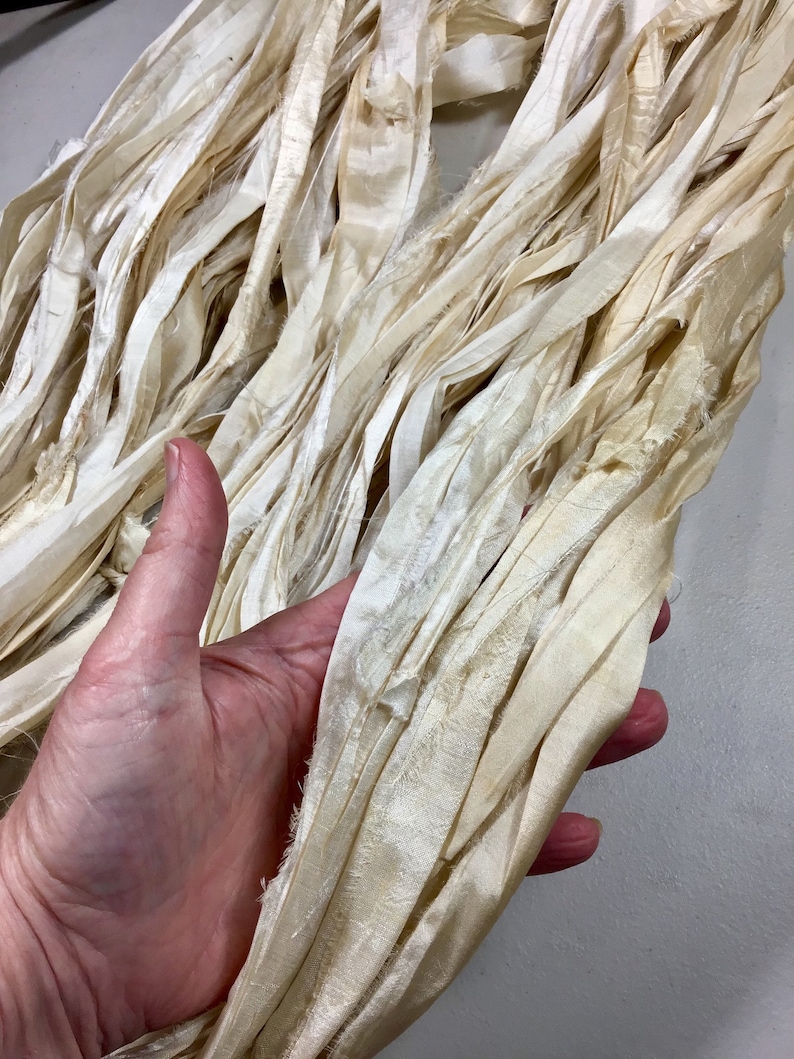 May include: Cream-colored silk ribbon, with a slightly frayed edge, is held in a hand. The ribbon has a soft, textured appearance and is arranged in loose folds. The color is a warm, off-white, suggesting a natural dye. This ribbon is suitable for crafts and embellishments.