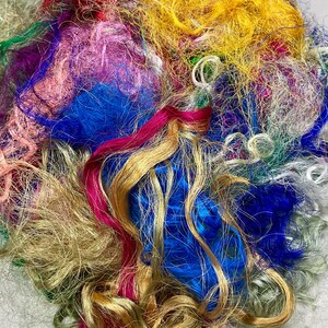 May include: A close-up of a pile of colorful craft fibers. The fibers are a mix of textures and colors, including gold, blue, pink, yellow, green, and red. The fibers appear to be made of silk or a similar material.