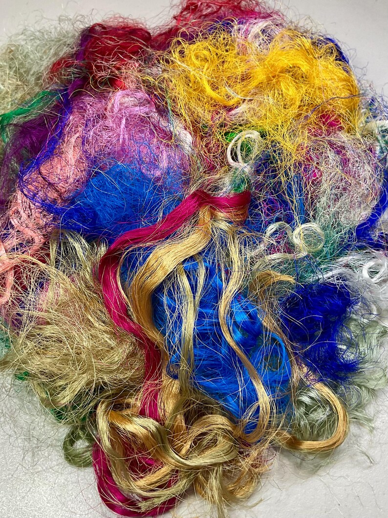 May include: A close-up of a pile of colorful craft fibers. The fibers are a mix of textures and colors, including gold, blue, pink, yellow, green, and red. The fibers appear to be made of silk or a similar material.