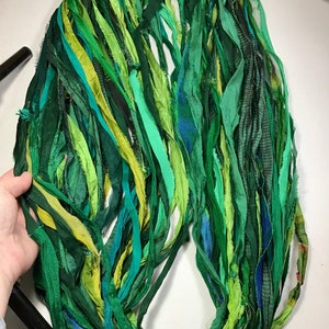 May include: A collection of green silk ribbon strips in various shades, including dark, medium, and light greens, with hints of yellow and blue. The ribbons are arranged in a circular shape, suggesting a craft supply or decorative element.