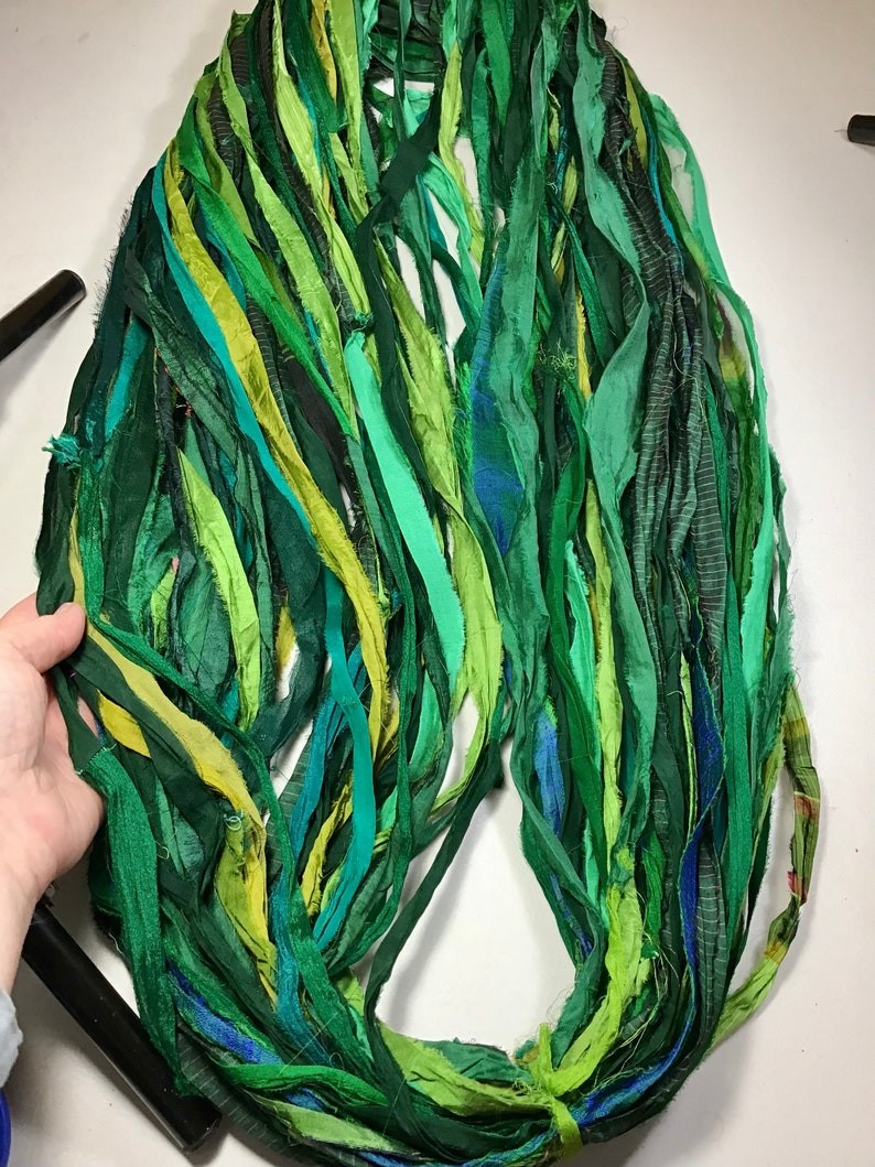 May include: A collection of green silk ribbon strips in various shades, including dark, medium, and light greens, with hints of yellow and blue. The ribbons are arranged in a circular shape, suggesting a craft supply or decorative element.