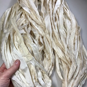 May include: A bundle of ivory-colored silk ribbon, with a slightly frayed texture. The ribbons are tightly bundled together, creating a dense, textured appearance. The silk has a subtle sheen, and the color varies slightly throughout the bundle.