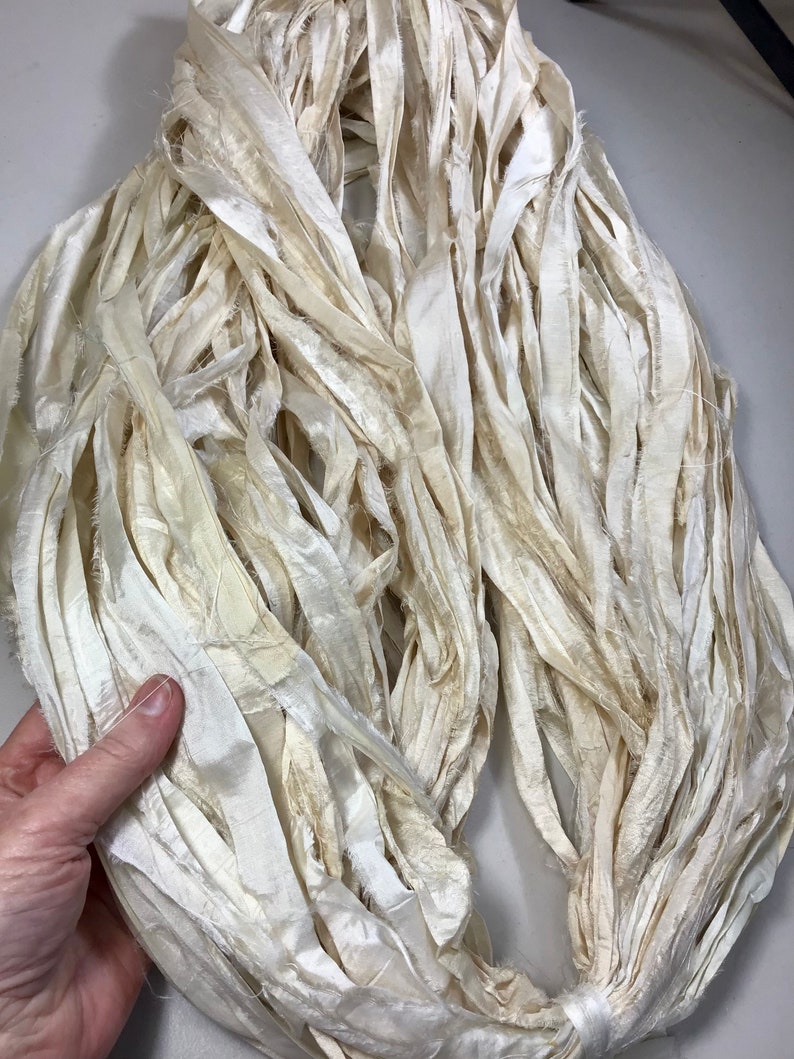 May include: A bundle of ivory-colored silk ribbon, with a slightly frayed texture. The ribbons are tightly bundled together, creating a dense, textured appearance. The silk has a subtle sheen, and the color varies slightly throughout the bundle.