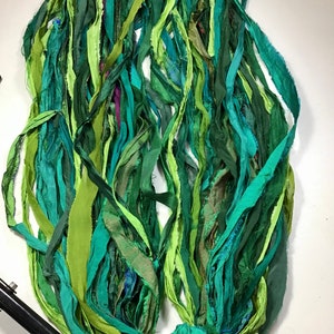 May include: A bundle of green fabric strips in various shades, including teal, lime, and forest green. The strips appear to be made of a soft, possibly silk, material. The fabric is arranged in a loose, flowing manner, suggesting it could be used for crafting or embellishment.