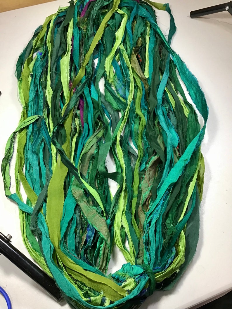 May include: A bundle of green fabric strips in various shades, including teal, lime, and forest green. The strips appear to be made of a soft, possibly silk, material. The fabric is arranged in a loose, flowing manner, suggesting it could be used for crafting or embellishment.