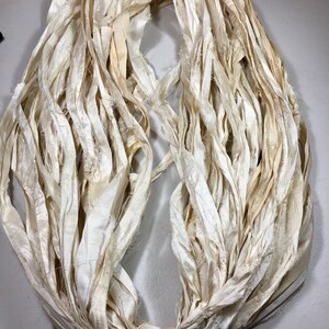 May include: A bundle of off-white, cream-colored silk ribbon. The ribbon is loosely arranged in a circular shape, with the ends tied together. The silk has a slightly distressed appearance, with some areas showing subtle discoloration.