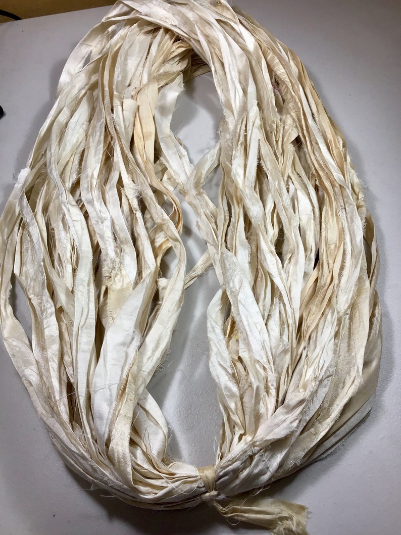 May include: A bundle of off-white, cream-colored silk ribbon. The ribbon is loosely arranged in a circular shape, with the ends tied together. The silk has a slightly distressed appearance, with some areas showing subtle discoloration.