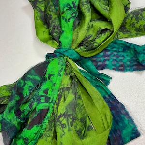 May include: A collection of green fabric strips tied together. The fabric features various shades of green, with some sections displaying a darker, patterned design. The strips are arranged in a layered, textured composition.