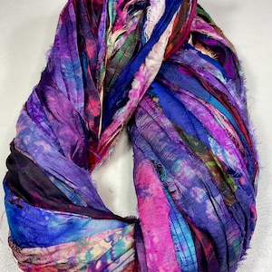 May include: A vibrant, hand-dyed silk scarf in shades of purple, blue, and pink. The scarf is made of strips of fabric, creating a textured, layered effect. The scarf is twisted into a circular shape.