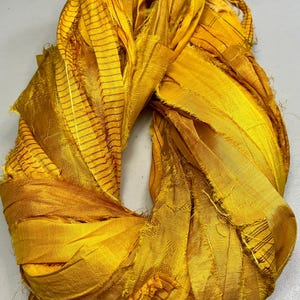 May include: Close-up of a bundle of golden-yellow silk ribbon. The ribbon is twisted and folded, revealing layers of textured fabric. Some sections have thin, dark brown stripes, adding visual interest. The overall effect is rich and luxurious.