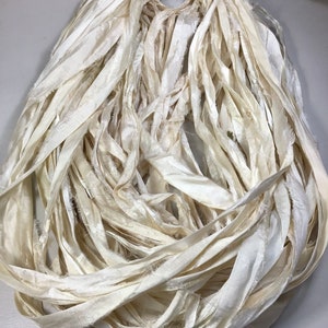 May include: A bundle of ivory-colored silk ribbon, tightly gathered at the top and cascading downwards. The ribbon has a slightly frayed texture, adding a rustic and handmade feel. Ideal for crafting and embellishments.