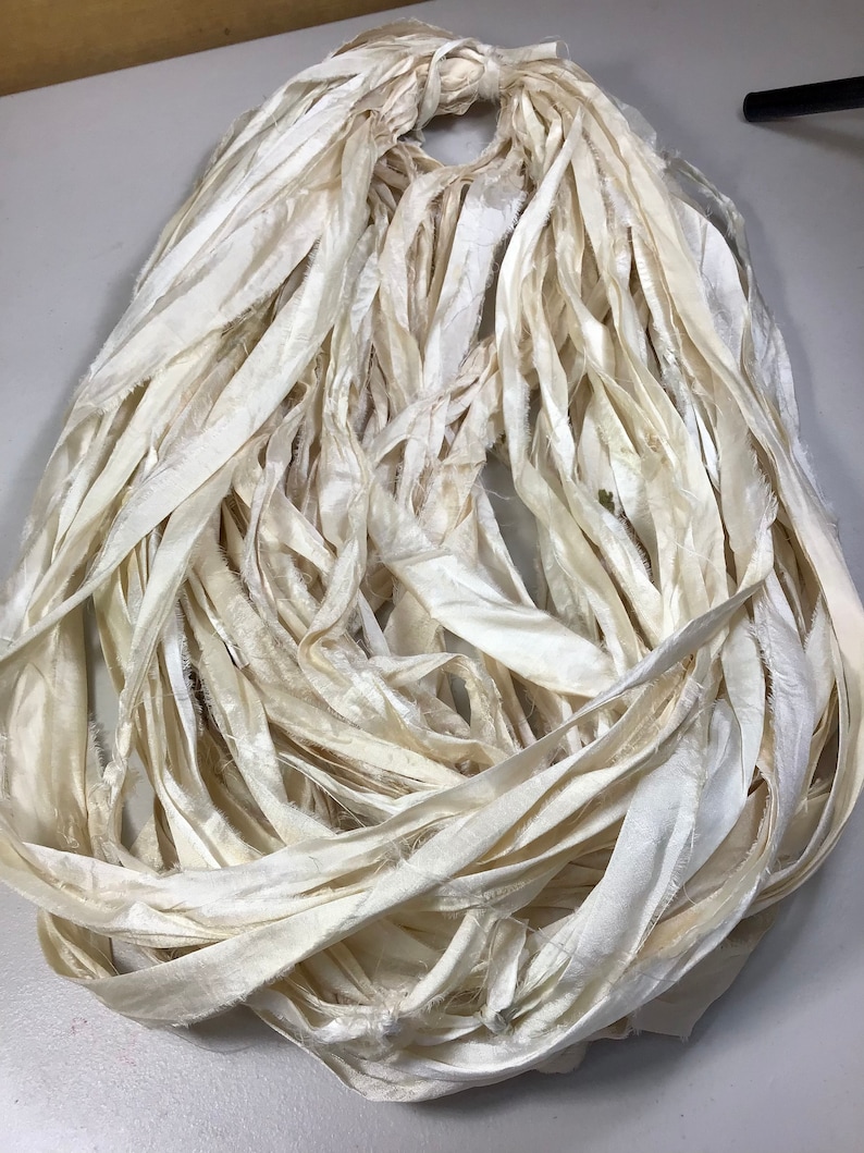 May include: A bundle of ivory-colored silk ribbon, tightly gathered at the top and cascading downwards. The ribbon has a slightly frayed texture, adding a rustic and handmade feel. Ideal for crafting and embellishments.