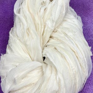 May include: A bundle of off-white silk ribbon featuring a crinkled, textured surface with raw, frayed edges. The ribbon is gathered into a loose, voluminous coil, showcasing the delicate, semi-sheer quality of the fabric and its soft, wavy folds.