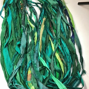 May include: A bundle of green silk ribbon strips in various shades, including teal, emerald, and olive. The ribbons are of varying widths and textures, creating a layered effect. The ribbons are bunched together at the top.