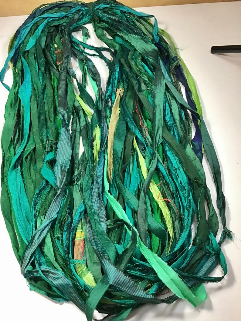 May include: A bundle of green silk ribbon strips in various shades, including teal, emerald, and olive. The ribbons are of varying widths and textures, creating a layered effect. The ribbons are bunched together at the top.