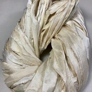 May include: A circular bundle of ivory-colored fabric strips. The fabric appears to be silk or a similar material, with a slightly textured surface and frayed edges. The strips are tightly wound, creating a dense, looped shape.