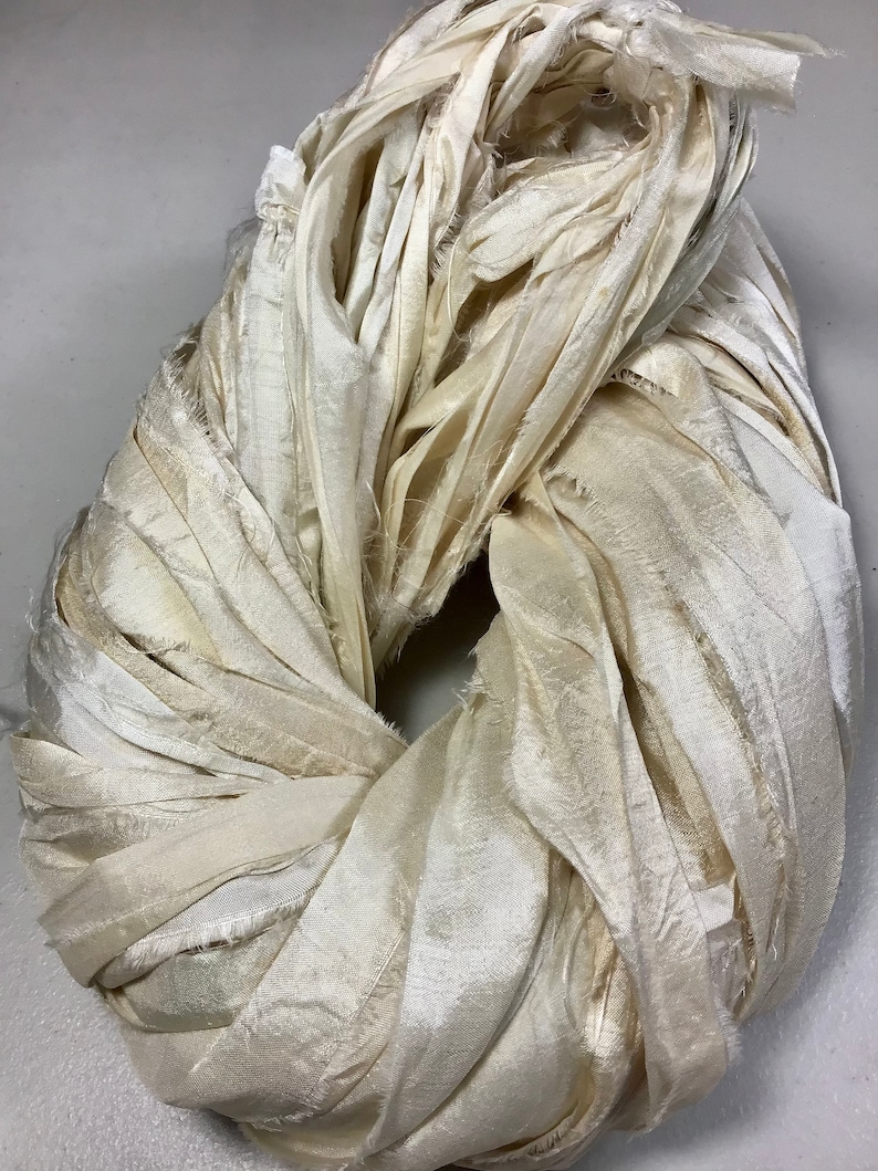 May include: A circular bundle of ivory-colored fabric strips. The fabric appears to be silk or a similar material, with a slightly textured surface and frayed edges. The strips are tightly wound, creating a dense, looped shape.