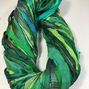 May include: A handmade wreath composed of various shades of green fabric strips. The strips are twisted and arranged in a circular shape, creating a textured and layered appearance. The wreath is suitable for home decor.