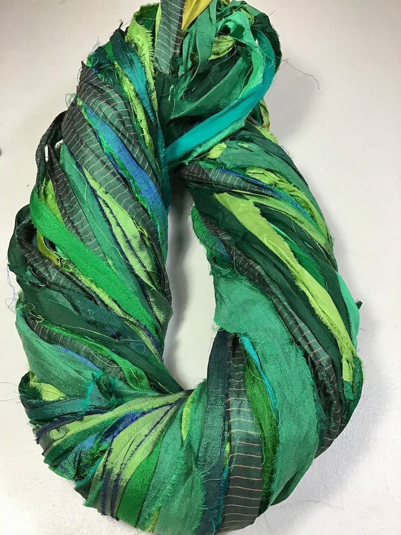 May include: A handmade wreath composed of various shades of green fabric strips. The strips are twisted and arranged in a circular shape, creating a textured and layered appearance. The wreath is suitable for home decor.