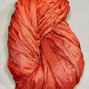 May include: A twisted bundle of vibrant orange silk ribbon. The ribbon has a textured, slightly frayed appearance, with visible threads and a rich, saturated color. The material appears soft and pliable, suggesting it could be used for crafting or decorative purposes.