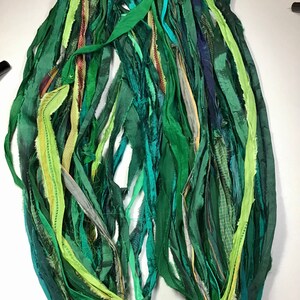 May include: A collection of green fabric strips in various shades, including emerald, forest, and lime. The strips are of varying widths and textures, some with subtle patterns. The arrangement suggests a craft supply or textile art material.