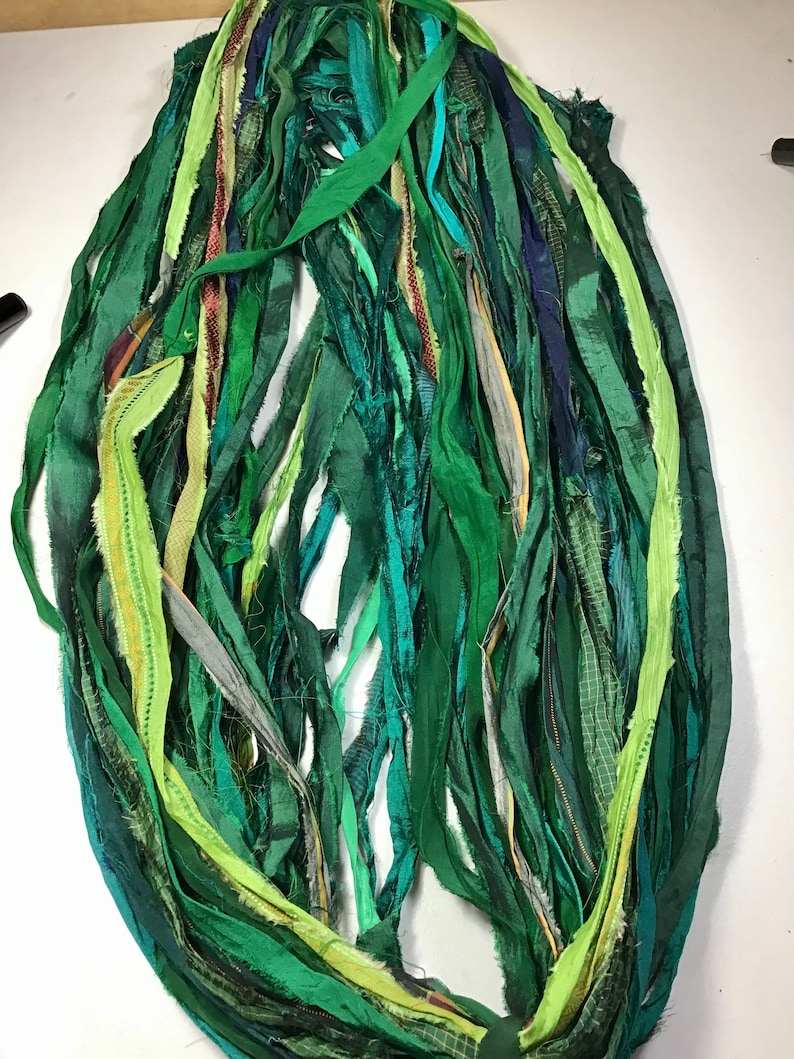 May include: A collection of green fabric strips in various shades, including emerald, forest, and lime. The strips are of varying widths and textures, some with subtle patterns. The arrangement suggests a craft supply or textile art material.
