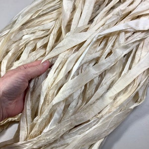 May include: Cream-colored, textured silk ribbon in a bundle. The ribbon is made of long, thin strips with frayed edges, creating a soft, flowing appearance. The ribbon is held by a hand, showing the texture and color variations.