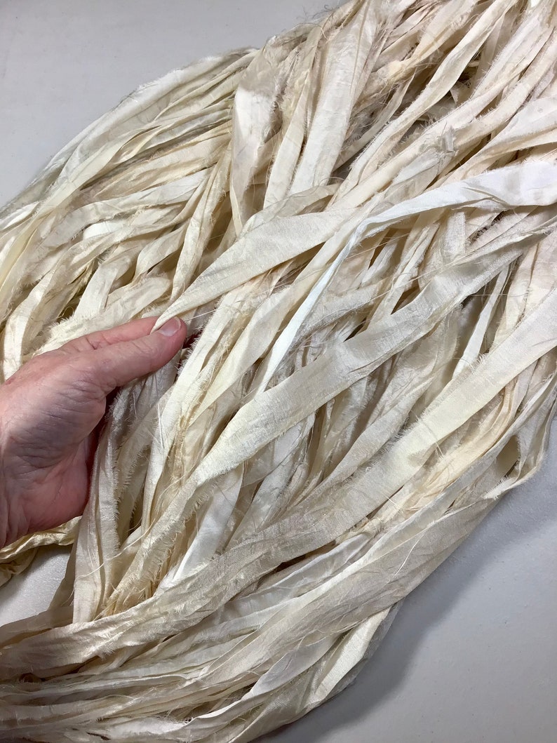 May include: Cream-colored, textured silk ribbon in a bundle. The ribbon is made of long, thin strips with frayed edges, creating a soft, flowing appearance. The ribbon is held by a hand, showing the texture and color variations.