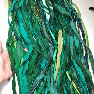 May include: A bundle of green fabric strips in various shades, including teal, emerald, and olive. The strips are of varying widths and textures, creating a layered effect. The fabric appears to be silk or a similar material, suitable for crafting or textile projects.