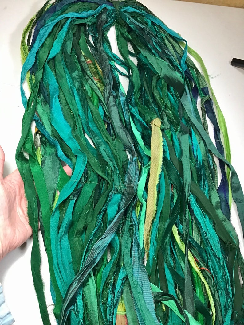 May include: A bundle of green fabric strips in various shades, including teal, emerald, and olive. The strips are of varying widths and textures, creating a layered effect. The fabric appears to be silk or a similar material, suitable for crafting or textile projects.
