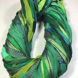 May include: A handmade wreath composed of various shades of green fabric strips. The strips are arranged in a circular shape, with a mix of textures and patterns, including solid, striped, and subtly textured fabrics. The wreath is a vibrant, decorative item.