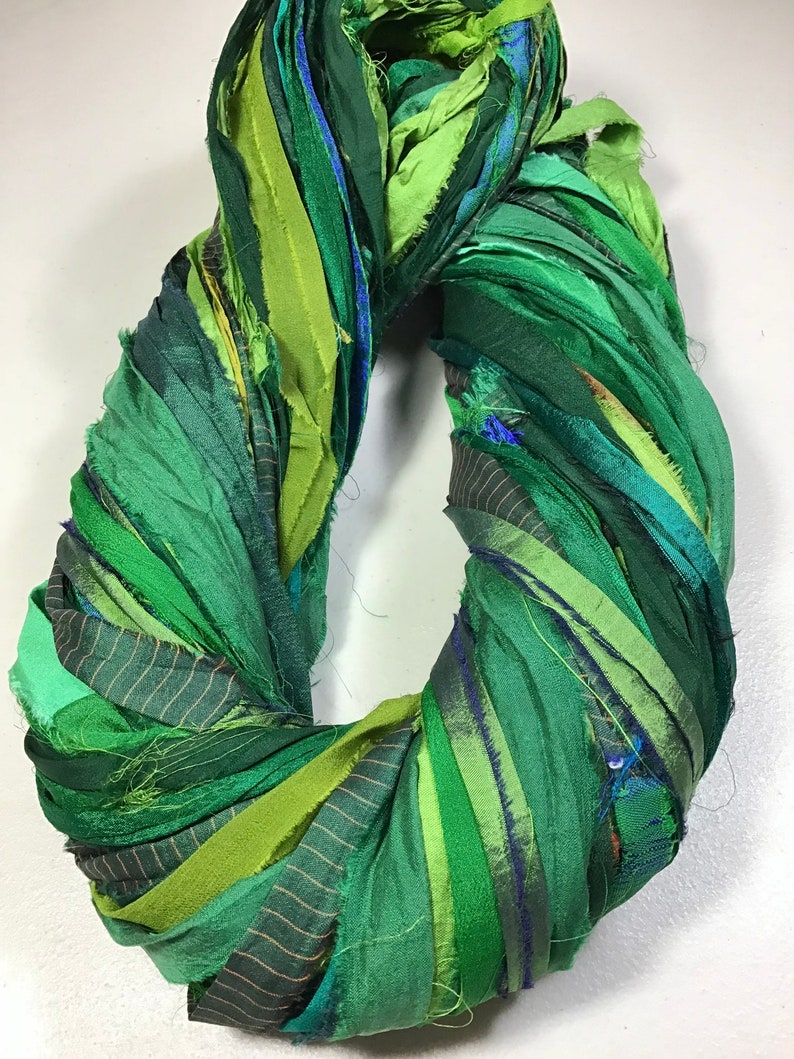 May include: A handmade wreath composed of various shades of green fabric strips. The strips are arranged in a circular shape, with a mix of textures and patterns, including solid, striped, and subtly textured fabrics. The wreath is a vibrant, decorative item.