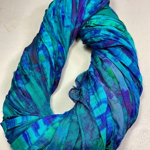 May include: A vibrant, hand-dyed silk scarf in shades of teal, turquoise, and deep blue. The scarf is made of strips of fabric, creating a textured, layered effect. The scarf is twisted into a circular shape.