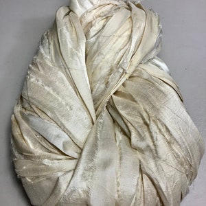 May include: A bundle of off-white fabric, possibly silk or satin, is tightly gathered and knotted. The fabric has a subtle sheen and visible texture, with frayed edges adding a raw, unfinished quality. The overall shape is rounded and compact.