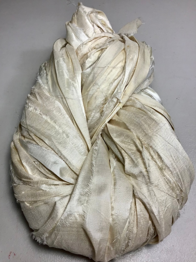 May include: A bundle of off-white fabric, possibly silk or satin, is tightly gathered and knotted. The fabric has a subtle sheen and visible texture, with frayed edges adding a raw, unfinished quality. The overall shape is rounded and compact.