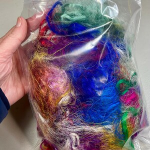 May include: A clear plastic bag filled with colorful strands of fiber in shades of blue, green, purple, red, and gold. The fibers appear to be synthetic and are tightly packed together. The bag is held by a hand.