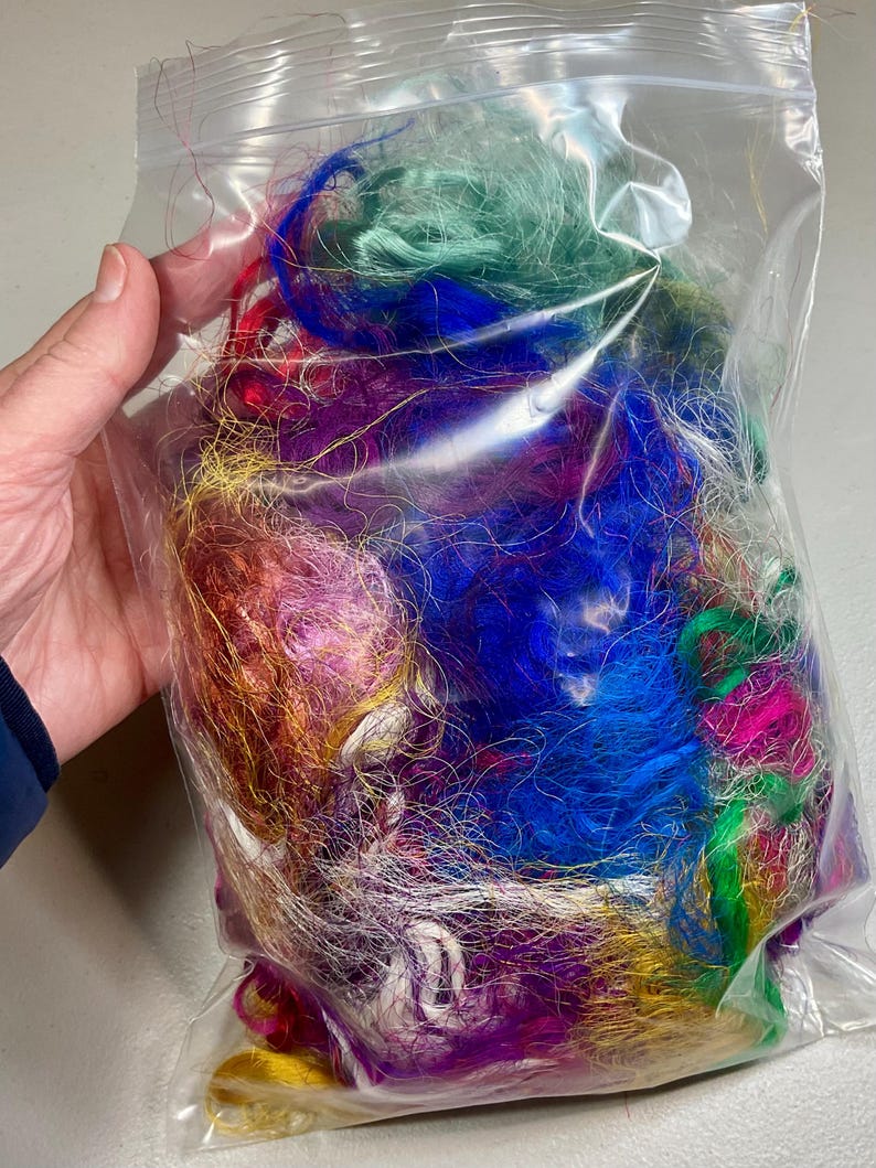 May include: A clear plastic bag filled with colorful strands of fiber in shades of blue, green, purple, red, and gold. The fibers appear to be synthetic and are tightly packed together. The bag is held by a hand.